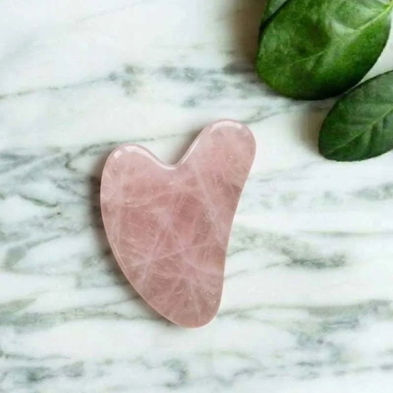 Gua Sha en quartz rose en forme de cœur posé sur du marbre, avec des feuilles vertes