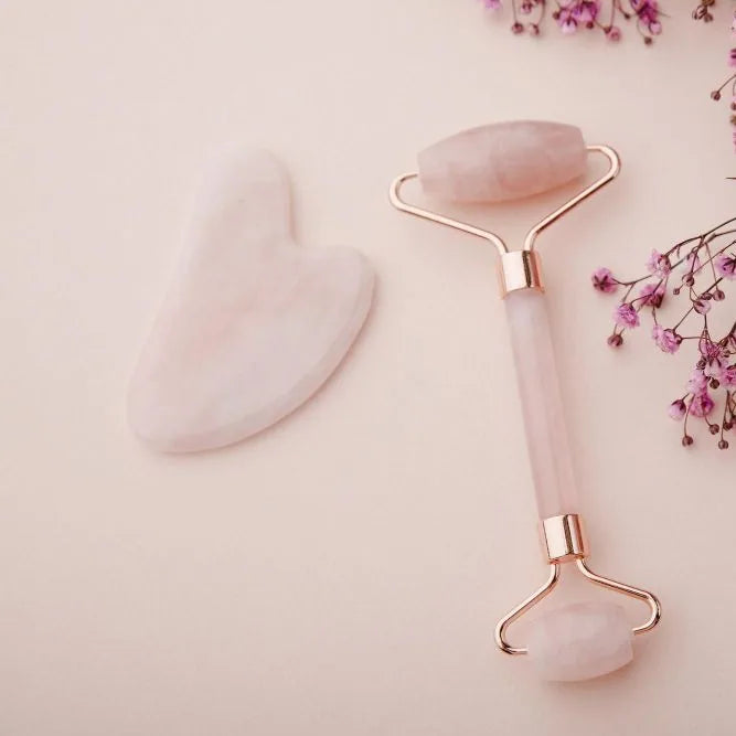 Kit rouleau et Gua Sha en quartz rose posés sur un fond rose pâle avec des fleurs